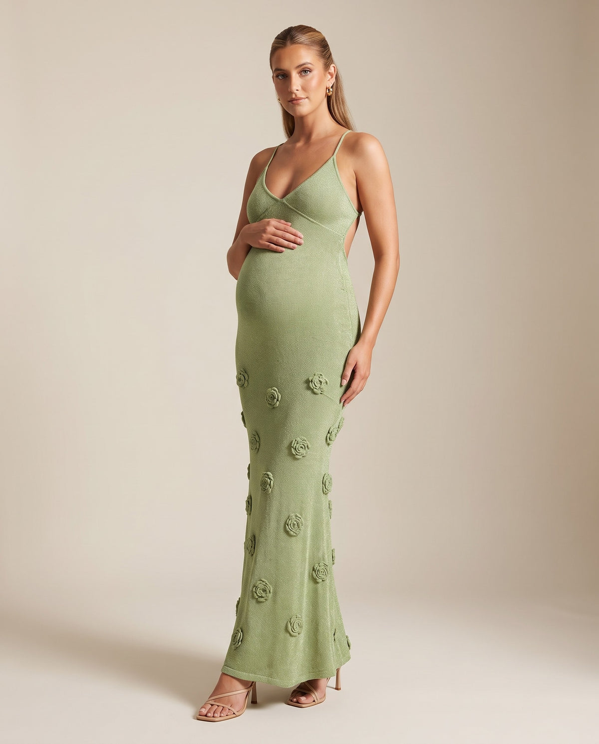 Sage green shimmer knit maternity maxi dress with 3D flowers, cami straps and open tie back, shown on a pregnant model in studio.