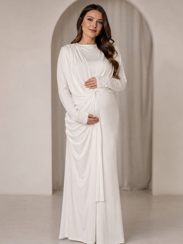 White jersey maternity maxi dress with draped cape overlay and long sleeves, designed for maternity photoshoot.