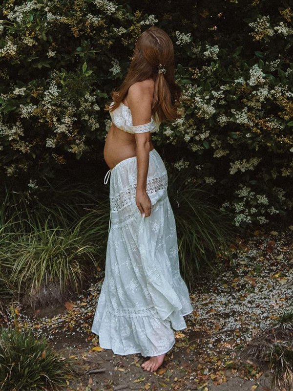 Back view of white eyelet embroidered maternity two-piece set with tie-front crop top and tiered lace-trim maxi skirt, designed for maternity photoshoot.