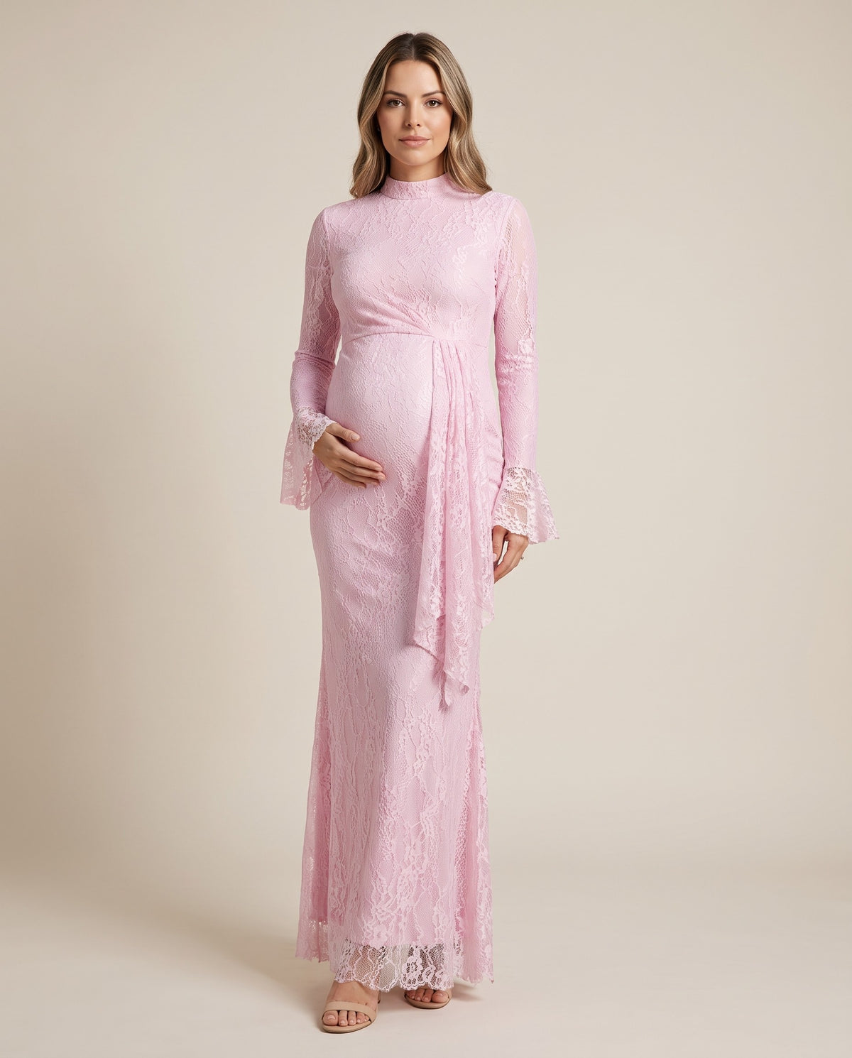Pink lace maternity maxi dress with high neck, bell sleeves and draped lace panel, shown on a pregnant model in studio.
