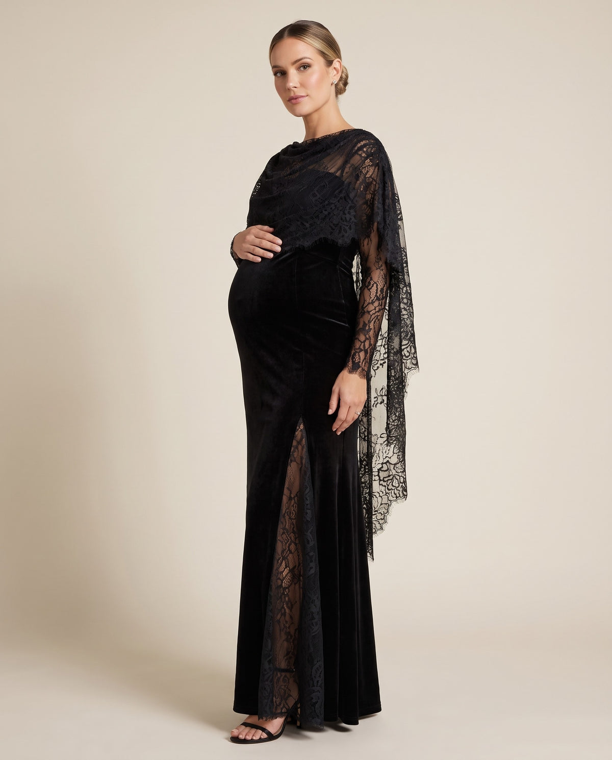Black velvet maternity maxi dress with sheer floral lace cape panel and lace sleeves, shown on a pregnant model in studio.